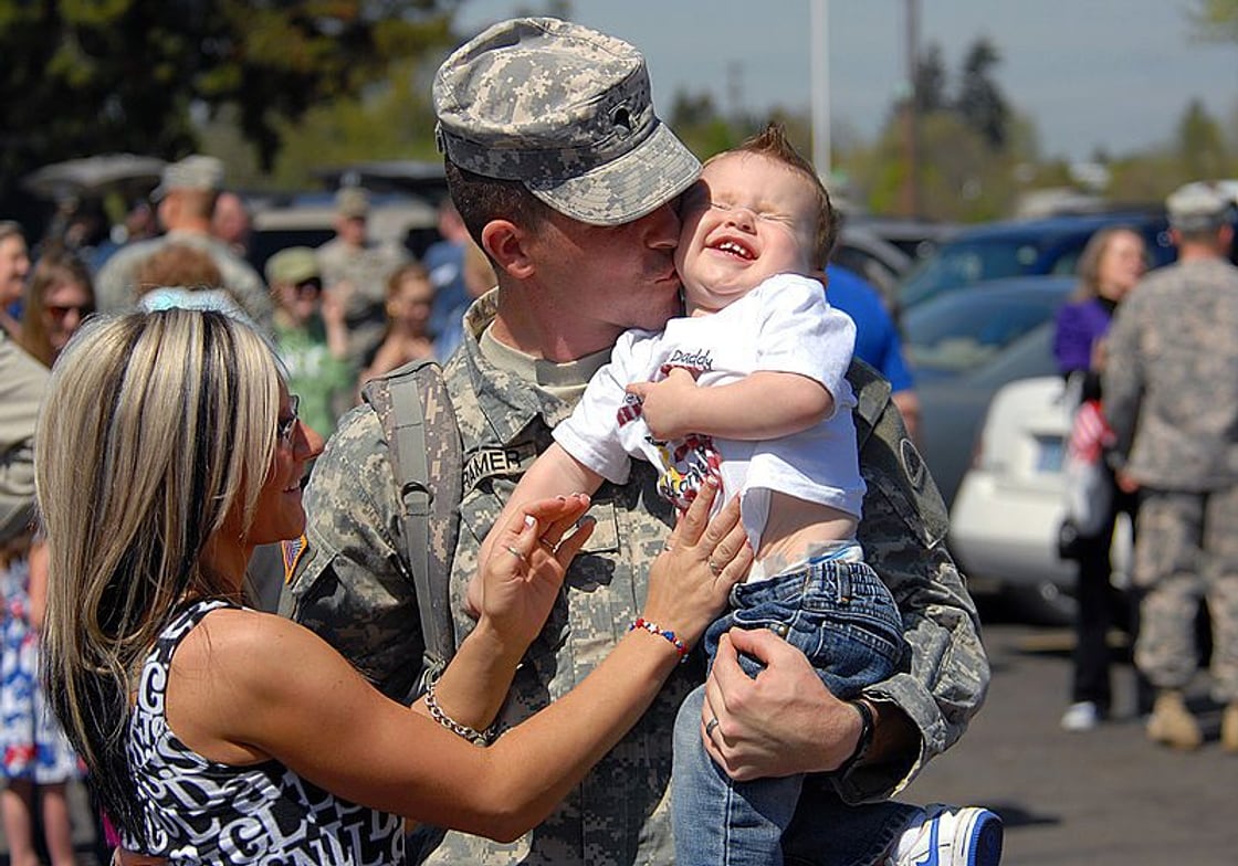 Soldier in uniform kissing his laughing toddler at a homecoming, his wife beside them